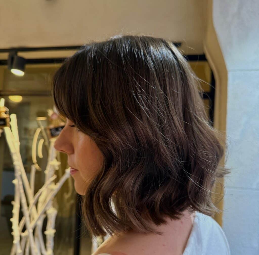 Femme de profil arborant un carré brun ondulé avec frange rideau, une coiffure moderne et texturée.