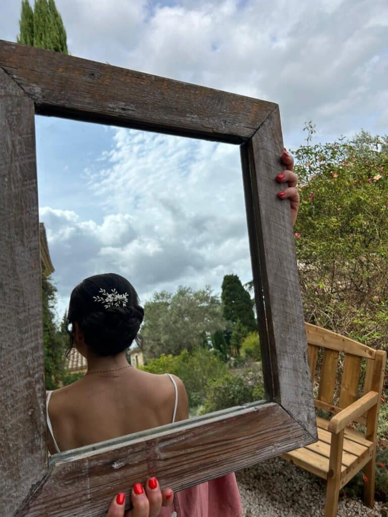 Reflet d'une femme avec chignon et accessoire floral dans un miroir rustique en bois tenu par des mains aux ongles rouges.